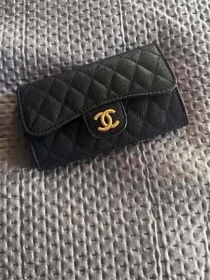 Svart quiltad väska  - Snygg svart plånbok från  i quiltat läder med klassiskt guldigt CC-spänne framtill. Plånboken har en tidlös design och känns lyxig med sin mjuka struktur och eleganta form. Perfekt för dig som gillar exklusiva accessoarer.