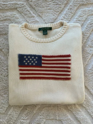 Vit stickad tröja Lauren Ralph Lauren - Vit stickad tröja från Lauren Ralph Lauren med amerikansk flagga broderad på bröstet och RLL-broderi vid nederkanten. Rund halsringning och ribbade muddar vid ärmslut och nederkant. Perfekt för dig som gillar klassisk preppy stil. Är dam men passar även till herr om man har xs eller är lite mindre så att man kan ha S.