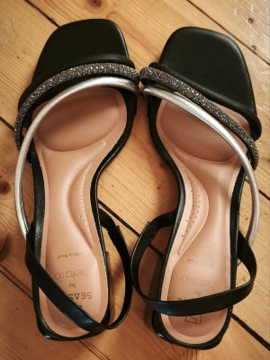 Svarta sandaler med glitter från Beira Rio - Sandaletter med remmar i svart och silverfärg, varav en rem är dekorerad med strass. Skorna har en bekväm innersula med dämpning fram och en stabil blockklack.  Använda vid två tillfällen, passar en 38 (jag har 37.5 ) annars glider remmen av på hälen.