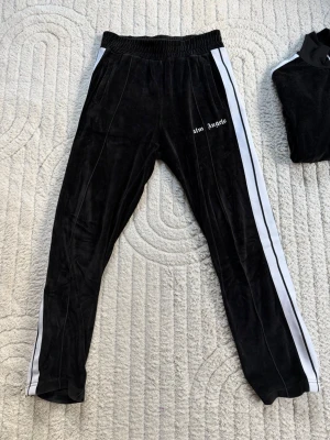 Svarta velourbyxor Palm Angels - Säljer ett par svarta mjukisbyxor från Palm Angels i mjuk velour med logotyp broderad på vänster sida. Byxorna har rak passform, resår i midjan och diskreta sömdetaljer framtill. Perfekta för en chill och trendig look.