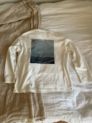 Vit långärmad t-shirt med tryck - Vit långärmad t-shirt med rund halsringning. På bröstet står det 'LEISURELY SEIZE THE DAY' och på ryggen finns ett tryck med havsmotiv och fåglar. Tillverkad i mjuk bomull för en skön känsla.