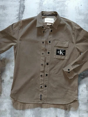 Dusty brown skjorta från Calvin Klein - Stilren dusty brun skjorta från Calvin Klein med svart logotyp-patch på bröstfickan. Skjortan har klassisk krage, långa ärmar med manschettknappar och svarta knappar framtill. Tillverkad i ett mjukt bomullstyg som ger en avslappnad men snygg look.