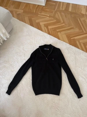 Svart stickad tröja från Polo Ralph Lauren - Säljer en svart stickad tröja från Polo Ralph Lauren med halvzip och klassisk logga broderad på bröstet. Tröjan har ribbstickad krage och muddar, perfekt för en stilren look. Materialet är mjukt och bekvämt, och modellen är långärmad.