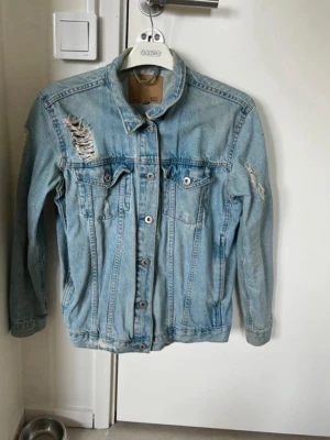 Ljusblå oversized jeansjacka - Säljer en ljusblå jeansjacka med slitna detaljer och broderi på ryggen. Jackan har två bröstfickor med knappar och klassisk krage. Perfekt för dig som gillar en avslappnad och trendig stil.