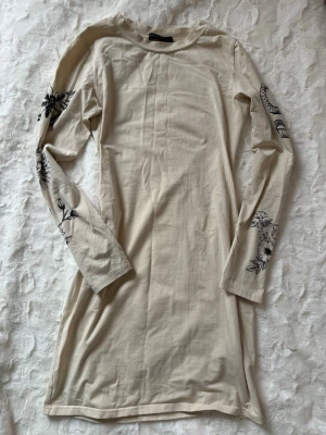 Beige långärmad klänning med blommor - Säljer en beige långärmad klänning med svart blommigt mönster på ärmarna. 