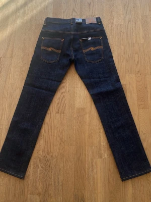 Mörkblå jeans från Nudie Jeans Thin Finn - Säljer ett par mörkblå jeans från Nudie Jeans med klassisk femficksdesign och orangea kontrastsömmar. Jeansen har raka ben och är tillverkade i kraftig denim. Snygga detaljer på bakfickorna med Nudie Jeans signaturbroderi.