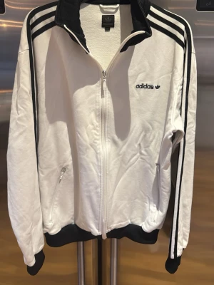 Vit Adidas zip-tröja med svarta detaljer - Vit zip-tröja från Adidas med svarta detaljer och klassiska tre ränder längs ärmarna. Tröjan har hög krage, två fickor med dragkedja och Adidas-logga på bröstet. Materialet är mjukt och bekvämt, perfekt för en sportig look. Vintage.
