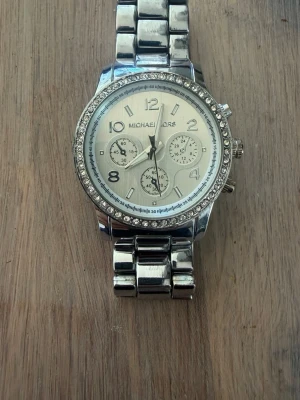  Michael Kors klocka med kristaller - Snygg silverfärgad klocka från Michael Kors, modell MK-5076. Klockan har ett klassiskt armband i rostfritt stål och en urtavla med tre små sub-dials. Ringen runt urtavlan är dekorerad med gnistrande kristaller för extra stil.