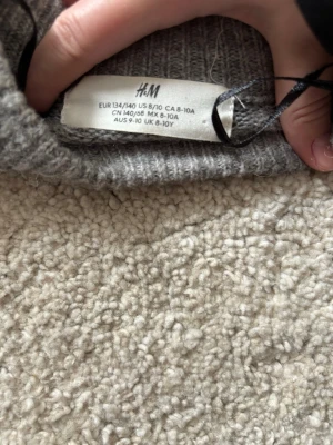 Grå stickad tröja med pärlor H&M - En grå stickad tröja från H&M med rund hals och långa ärmar. Tröjan har dekorativa pärlor på framsidan och ribbstickade muddar vid ärmslut och nederkant. Perfekt för att ge din outfit en extra touch.