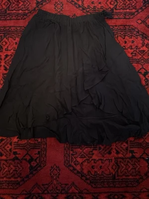 Svart omlottkjol i polyester - Svart långkjol med omlottdetalj och elastisk midja. Kjolen är tillverkad i lätt polyester och har en enkel, flowy design som passar dig som gillar stilrena plagg. Perfekt att matcha med sneakers eller boots.