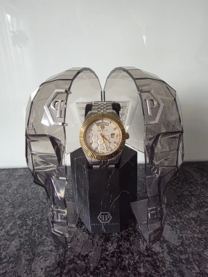 Philipp Plein herrklocka i silver och guld - Säljer en Philipp Plein herrklocka med silverfärgad länkarmband och guldfärgad boett. Urtavlan är vit med hexagonmönster, dag- och datumvisning samt tydliga guldfärgade visare. Klockan levereras i en unik display med två genomskinliga dödskallar.