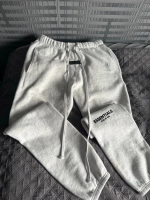 Grå Essentials mjukisbyxor - Säljer ett par grå mjukisbyxor från Essentials med logga på vänster ben. Byxorna har snörning i midjan och resår vid benslut. Perfekta för chill och avslappnade dagar, tillverkade i mjukt bomullsmaterial.