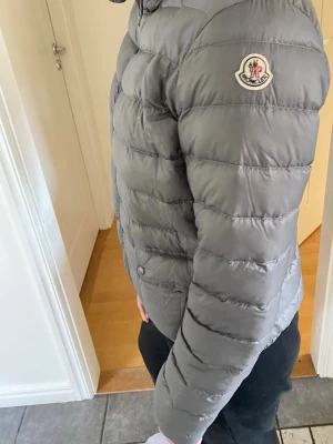 Grå dunjacka från Moncler - Säljer en grå dunjacka från Moncler med klassisk quiltad design och två fickor med knappstängning framtill. Jackan har hög krage och Moncler-logga på ärmen. Perfekt för kalla vinterdagar och riktigt snygg att bära till jeans eller joggers.