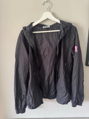 Svart vindjacka från Risky Universe - Svart vindjacka från Risky med huva och dragkedja. Jackan har rosa tryck på ärmen och ett stort rosa Risky-tryck med handmotiv på huvan. Tillverkad i lätt, prassligt material som passar perfekt för blåsiga dagar. Justerbar nederkant med snören. Storlek M