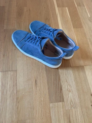 Blå mockasneakers från Christian Louboutin - Snygga blå sneakers i mocka från Christian Louboutin med klassisk röd sula och vita kanter. Skorna har lågt skaft, matchande blå skosnören och en stilren design som sticker ut med den ikoniska röda undersidan.