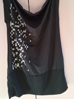 Svart ärmlös blus med paljetter - Snygg svart ärmlös blus med draperad urringning och silvriga paljettdetaljer på ena sidan. Blusen har en lös passform och är tillverkad i ett glansigt, mjukt material som ger ett elegant intryck. Perfekt för dig som vill sticka ut lite extra.