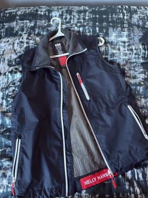 Svart Helly Hansen väst med dragkedjor - Cool svart väst från Helly Hansen med vita och röda detaljer. Västen har flera dragkedjor och meshfoder på insidan. Perfekt för lager-på-lager och har en sportig look med logotypen synlig på insidan.