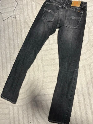 Grå jeans från Nudie Jeans - Säljer ett par grå jeans från Nudie Jeans med raka ben och klassisk femficksmodell. Jeansen har vita kontrastsömmar och Nudie-loggan på bakfickan samt läderpatch i midjan. Perfekta för en avslappnad och stilren look.