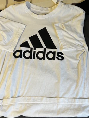 Vit Adidas t-shirt med logga - Vit t-shirt från Adidas med klassisk svart logga på bröstet. T-shirten har rund hals och korta ärmar, tillverkad i mjuk bomull som är skön att bära. Perfekt för dig som gillar sportiga och stilrena plagg.