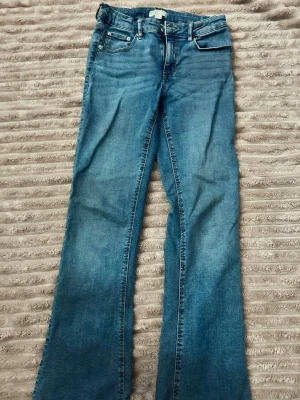 Blå bootcut jeans med fickdetaljer - Snygga blå bootcut jeans med klassisk femficksdesign och dekorativa ficklock bak. Jeansen har normalhög midja och är tillverkade i ett mjukt denimtyg. Perfekta för dig som gillar en avslappnad men trendig look.