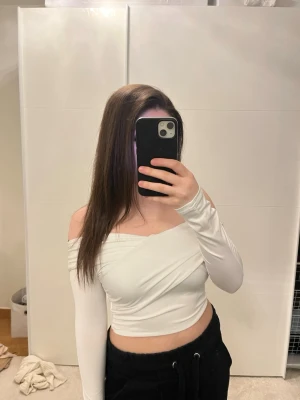 Vit offshoulder croppad topp - Trendig vit offshoulder topp med lång ärm och omlott-design. Croppad modell som sitter snyggt och markerar midjan. Perfekt för dig som vill ha en stilren och modern look.