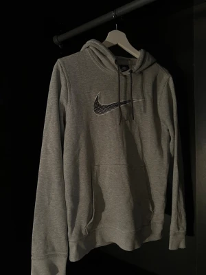 Grå Nike hoodie med logga - Snygg grå hoodie från Nike med stor svartvit Swoosh-logga på bröstet. Klassisk känguruficka framtill och justerbar huva med dragsko. Tillverkad i mjukt sweatshirtmaterial, perfekt för chill eller streetwear.