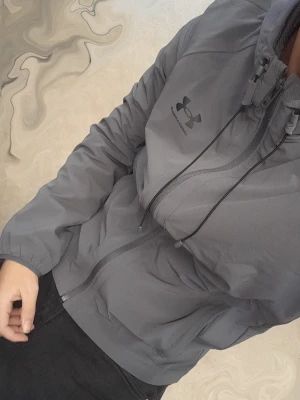 Grå vindjacka från Under Armour - Snygg grå vindjacka från Under Armour med dragkedja framtill och justerbar huva med snören. Jackan har elastiska muddar vid ärmsluten och logga på bröstet. Perfekt för blåsiga dagar och sportiga outfits.