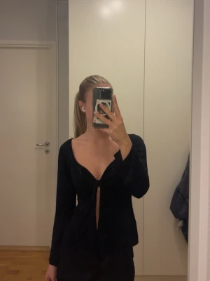 Svart långärmad topp med knyt - Snygg svart långärmad topp med djup v-ringning och knyt framtill. Toppen har en figurnära passform och är perfekt för dig som vill ha en trendig och stilren look. Materialet är mjukt och stretchigt.