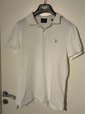 Vit pikétröja från Gant - Klassisk vit pikétröja från Gant med korta ärmar och liten logga broderad på bröstet. Tröjan har krage och knappar framtill, perfekt till en clean och stilren look. 