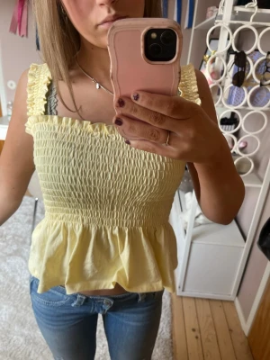 Gul smockad linnetopp med volang - Söt ljusgul linnetopp med smockad överdel och volang nedtill. Toppen är ärmlös med breda axelband och har en feminin, croppad passform. Perfekt för varma dagar och enkel att styla med jeans eller kjol.