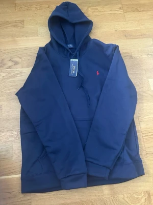Marinblå hoodie från Polo Ralph Lauren - Snygg marinblå hoodie från Polo Ralph Lauren med klassisk huva och dragsko. Ikoniska röda logotypen broderad på bröstet. Tillverkad i mjukt material och har en stor magficka framtill. Perfekt för en avslappnad och stilren look.