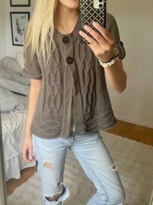 Brun kabelstickad kofta med knappar - Kortärmad brun kofta med kabelstickat mönster och stora dekorativa knappar framtill. Koftan har en avslappnad passform och ribbade detaljer vid ärmslut och nederkant. Perfekt att slänga över en t-shirt för en chill look. 