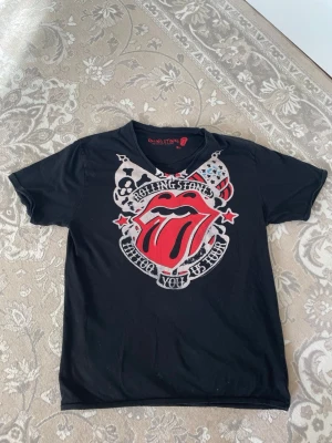 Rolling Stones svart t-shirt - Svart t-shirt med stort Rolling Stones-tryck framtill, klassiska röda tungan och texten 'Who do you love'. Rund hals och korta ärmar. Perfekt för dig som gillar bandtröjor och vill sticka ut med en ikonisk design.