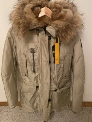 Parajumper jacka - 100% äkta. Snygg beige vinterjacka från Parajumpers med stor luva och fluffig pälskant. Jackan har flera fickor, dragkedja och knappar framtill samt den klassiska gula remmen både fram och bak. Perfekt för kalla dagar. Inga skador alls och inga tecken på användning. Passar XS och S. PRISET GÅR ATT DISKUTERA 