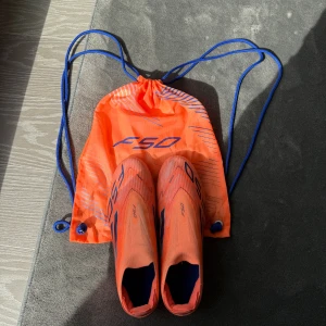 Adidas F50 orange fotbollsskor - Säljer ett par grymma Adidas F50 fotbollsskor i en riktigt snygg orange färg med blå detaljer. Skorna har låg profil, spetsig tå och är tillverkade i lätt syntetmaterial för maxad speed på planen. Medföljer matchande orange skopåse med blå snören. Köpta begagnande och använd en gång. 