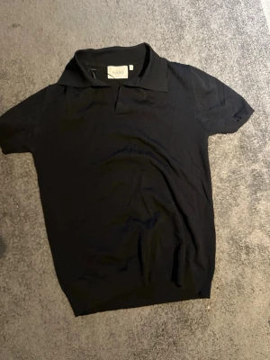 Svart pikétröja från Sandö - Klassisk svart pikétröja från Sandö med krage och korta ärmar. Tillverkad i mjukt material som ger en clean och stilren look. Perfekt till jeans eller chinos för en enkel men snygg outfit.