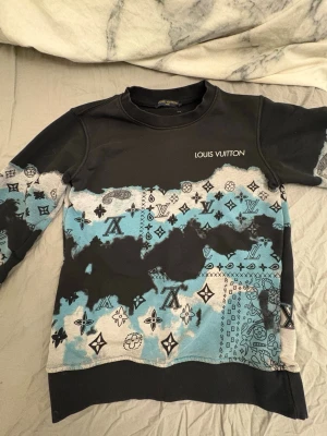 Louis Vuitton sweatshirt med blått mönster - Säljer en svart sweatshirt från Louis Vuitton med blått och vitt batikmönster samt klassiska LV-loggor och paisleydetaljer. Tröjan har rund halsringning och långa ärmar. Perfekt för dig som vill sticka ut med en unik designertröja.