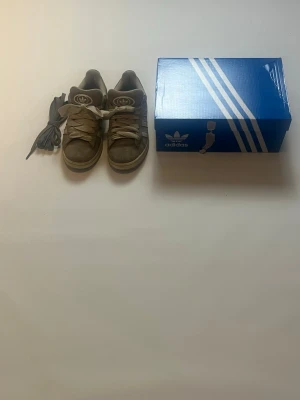Grå Adidas Campus 00 sneakers storlek 39 - Gråa Adidas Campus 00 sneakers i mocka med vita ränder på sidorna och vita skosnören. Klassisk låg modell med rund tå och vadderad plös. Skorna kommer med originalkartong och extra skosnören. Perfekta för en avslappnad streetstyle-look. De är också i storlek 39