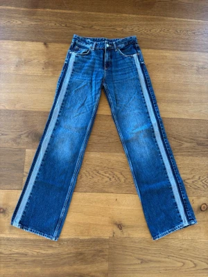 Blå jeans med kontrastpaneler - Säljer ett par blå jeans med raka ben och ljusare kontrastpaneler längs sidorna. Klassisk femficksmodell med bälteshällor och knappgylf. Jeansen har en cool vintagekänsla och är tillverkade i kraftigt denimtyg.