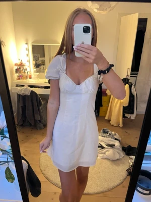 Vit kort klänning med puffärm - Söt vit kort klänning med puffärmar och markerad byst. Klänningen har en feminin siluett och är tillverkad i ett lätt tyg som ger en luftig känsla. Perfekt för sommarens alla tillfällen och enkel att styla med accessoarer.