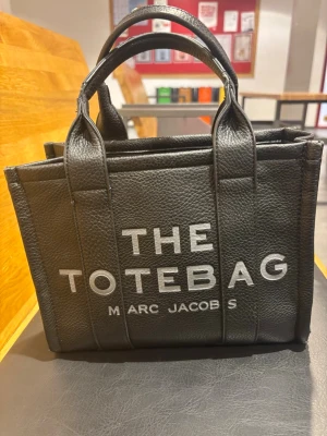 Svart The Tote Bag - Säljer en svart handväska, modell The Tote Bag. Väskan är tillverkad i strukturerat läder med två korta handtag och tydlig vit logga framtill. Klassisk boxig form som rymmer det viktigaste och passar perfekt till en trendig look. Lite slitage på handtagen. Säljer pga att den inte kommer till användning och säljer hellre vidare.