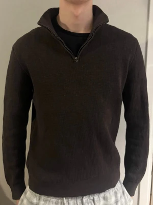 Mörkbrun stickad halvziptröja - Mörkbrun stickad tröja med halv dragkedja och hög krage. Tröjan har lång ärm och ribbade muddar vid ärmslut och nederkant. Perfekt för lager på lager och en clean look. Passar till både jeans och chinos.