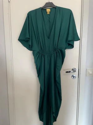 Grön långklänning med v-ringning - Snygg grön långklänning med djup v-ringning och vida korta ärmar. Klänningen har resår i midjan för en smickrande passform och ett mjukt, följsamt tyg som ger ett elegant fall. Perfekt för dig som vill ha en stilren och trendig look.