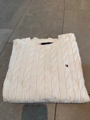 Vit kabelstickad tröja Tommy Hilfiger - Kabelstickad vit tröja från Tommy Hilfiger med rund halsringning och diskret logga broderad på bröstet.  Helt ny! Använd 2-3 gånger. Storlek 41.