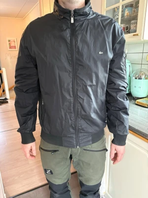 Svart vindjacka från Lacoste - Snygg svart vindjacka från Lacoste med hög krage och dragkedja framtill. Jackan har två fickor med dragkedja, ribbade muddar vid ärmslut och nederkant samt den klassiska Lacoste-loggan på bröstet. Tillverkad i lätt och tåligt material, perfekt för blåsiga dagar. Storlek passar som medium, person på bild är 180cm..