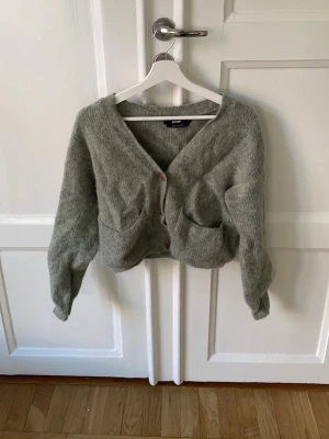 Grågrön kofta i alpaca blend från Bik Bok - Mysig grå-grön kofta från Bik Bok i alpaca blend. Koftan har v-ringning, stora framfickor och stängs med tre knappar framtill. Ej nopprig. 