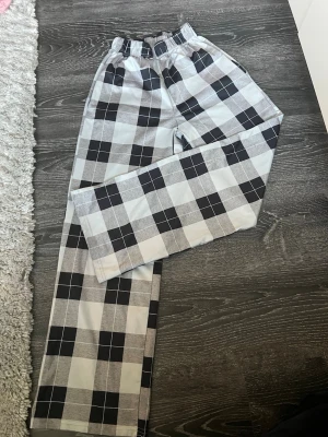 Rutiga mjukisbyxor i svart och vitt - Säljer ett par mjukisbyxor med stort rutmönster i svart, vitt och grått. Byxorna har resår i midjan och raka ben för en avslappnad look. Perfekta för chill dagar hemma eller när du vill vara bekväm men ändå stilren.