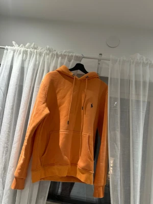 Orange hoodie  - Snygg orange hoodie med klassisk huva och dragsko. Tröjan har en känguruficka framtill och den ikoniska lilla loggan broderad på bröstet. Tillverkad i mjukt bomullsmaterial och har ribbade muddar vid ärmslut och nederkant.