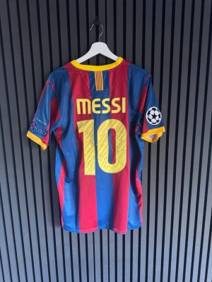 Barcelona Messi matchtröja Nike - Säljer en ikonisk FC Barcelona matchtröja med Messi och nummer 10 på ryggen. Tröjan är blå och röd med gula detaljer, officiella Champions League-märken på ärmen och klubbmärke på bröstet. Tillverkad i lätt och ventilerande material, perfekt för fotboll eller samling.