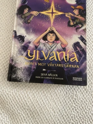 Ylvania: Hotet mot Väktarstjärnan - En spännande fantasybok med färgstarkt omslag i lila, gult och svart. Boken är skriven av Ylva Hällen och illustrerad av Marcus Gunnar Pettersson. Perfekt för dig som gillar magi, äventyr och mystiska världar. Inbunden med snyggt tryck och detaljer.